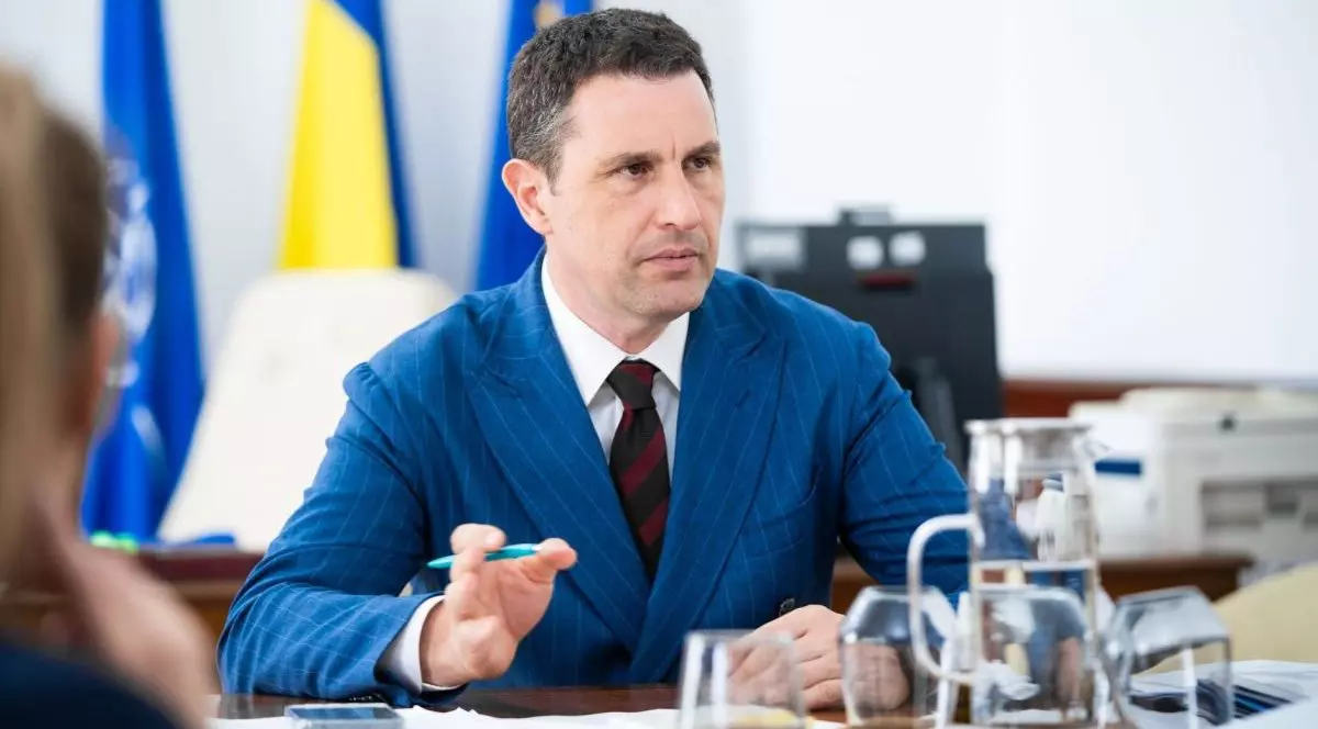 Ministrul interimar al Finanțelor, Tanczos Barna, îmbrăcat la costum, stă la o masă unde vorbește și explică dând din mâna stângă, în care are și un pix, iar în fața lui sunt pahare cu apă și în spate se văd steagul României, UE și al NATO