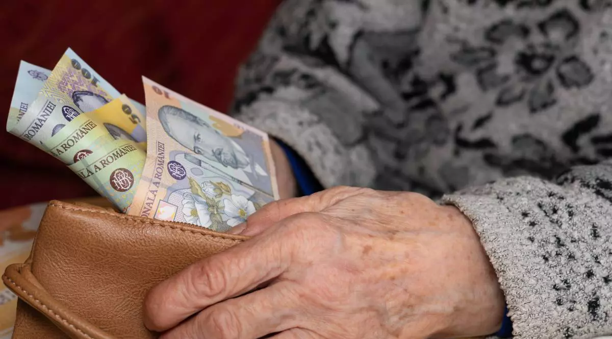 O femeie în vârstă care stă jos și își numără banii din portofel. Femeia poartă un pulover gri cu un model, portofelul pe care îl ține în mână este din piele maro, iar din portofel ies bancnote de 100, 50 și 10 lei.