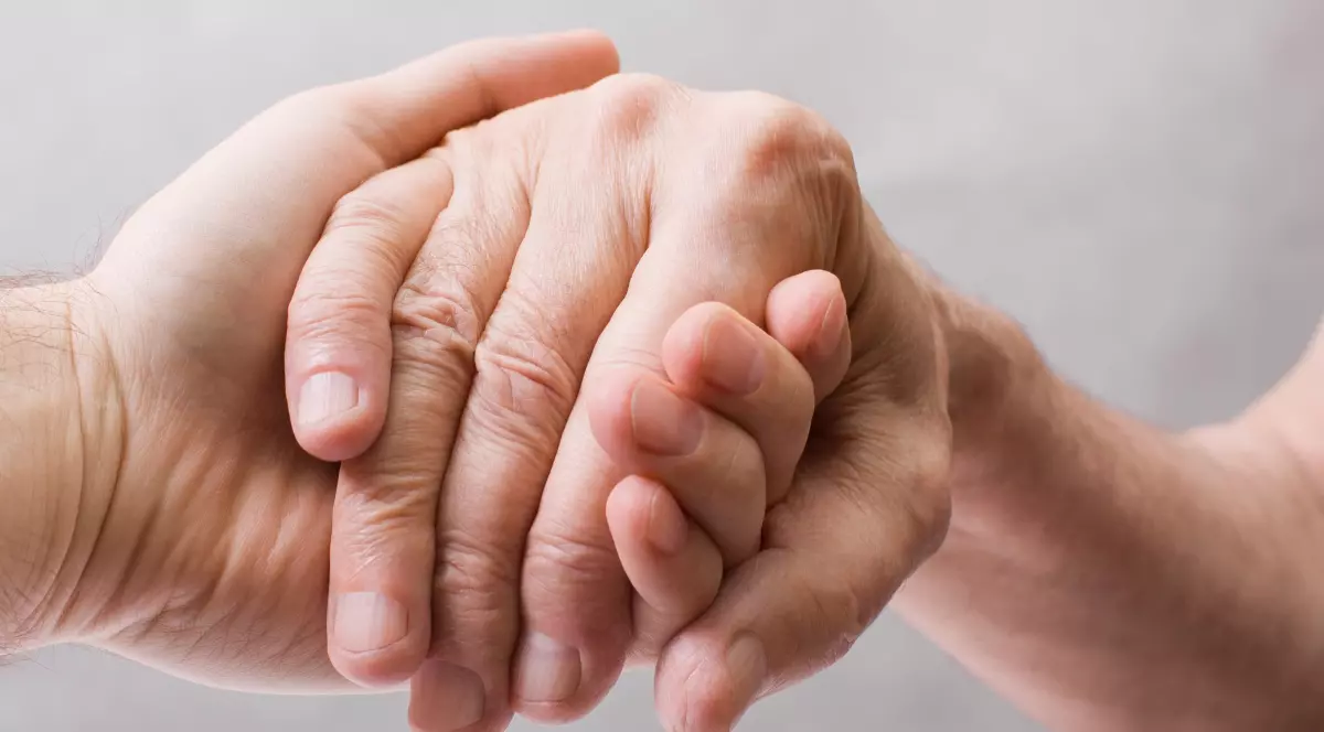 O persoană ține cu grijă mâna altei persoane între palmele sale. Mâna sprijinită pare a fi a unei persoane în vârstă