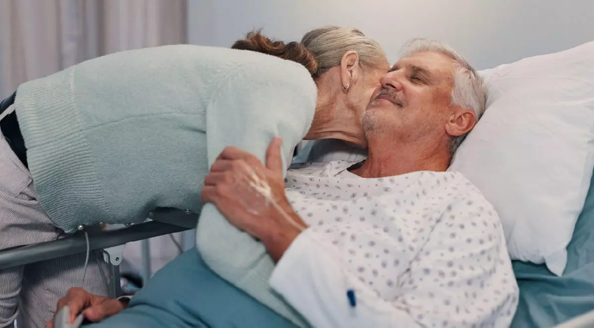 Femeie in varsta imbratisand un barbat cu parul alb, in pijama alba, pe un pat de spital
