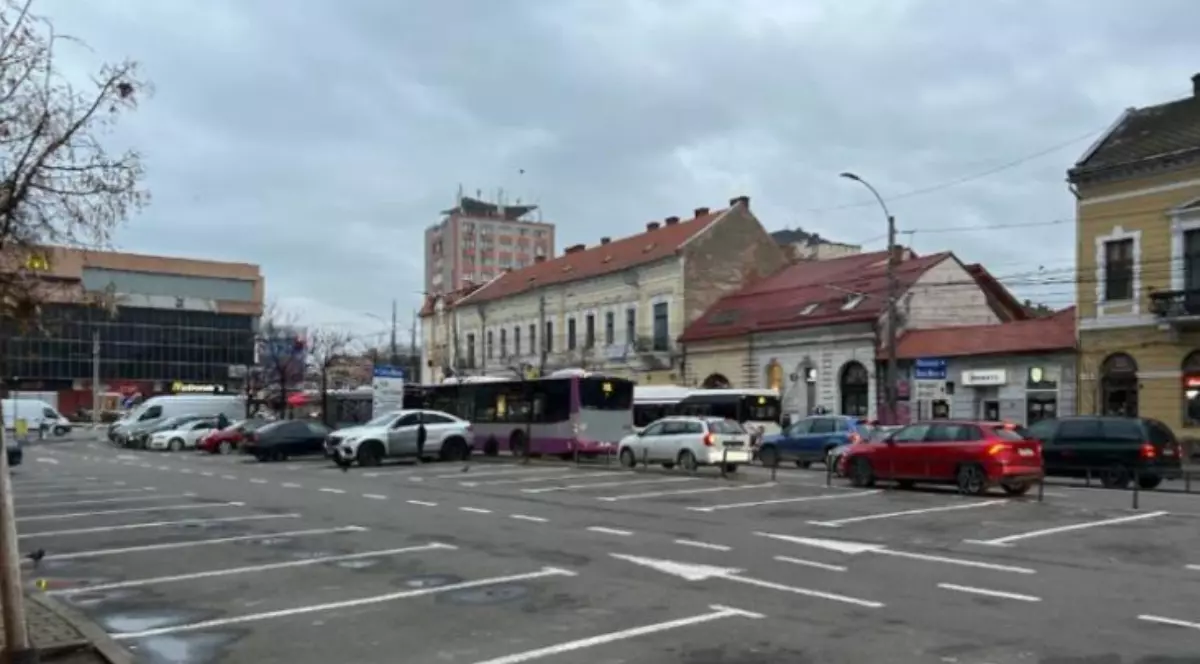 Imaginea surprinde parcarea din Piața Mihai Viteazul din Cluj-Napoca, într-o zi mohorâtă. Se pot observa mai multe mașini parcate, autobuze în stație.