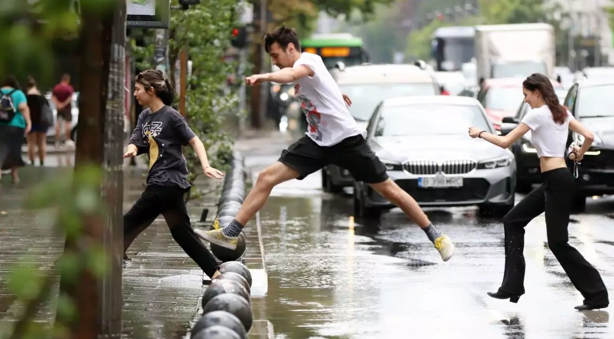 Un tânăr în tricou alb face un pas mare ca să traverseze strada, fiind o baltă de la ploaie, iar o fată în pantaloni lungi negri și cu tocuri, în spatele lui, încearcă să facă același lucru, într-o zi ploioasă, în București, 2025