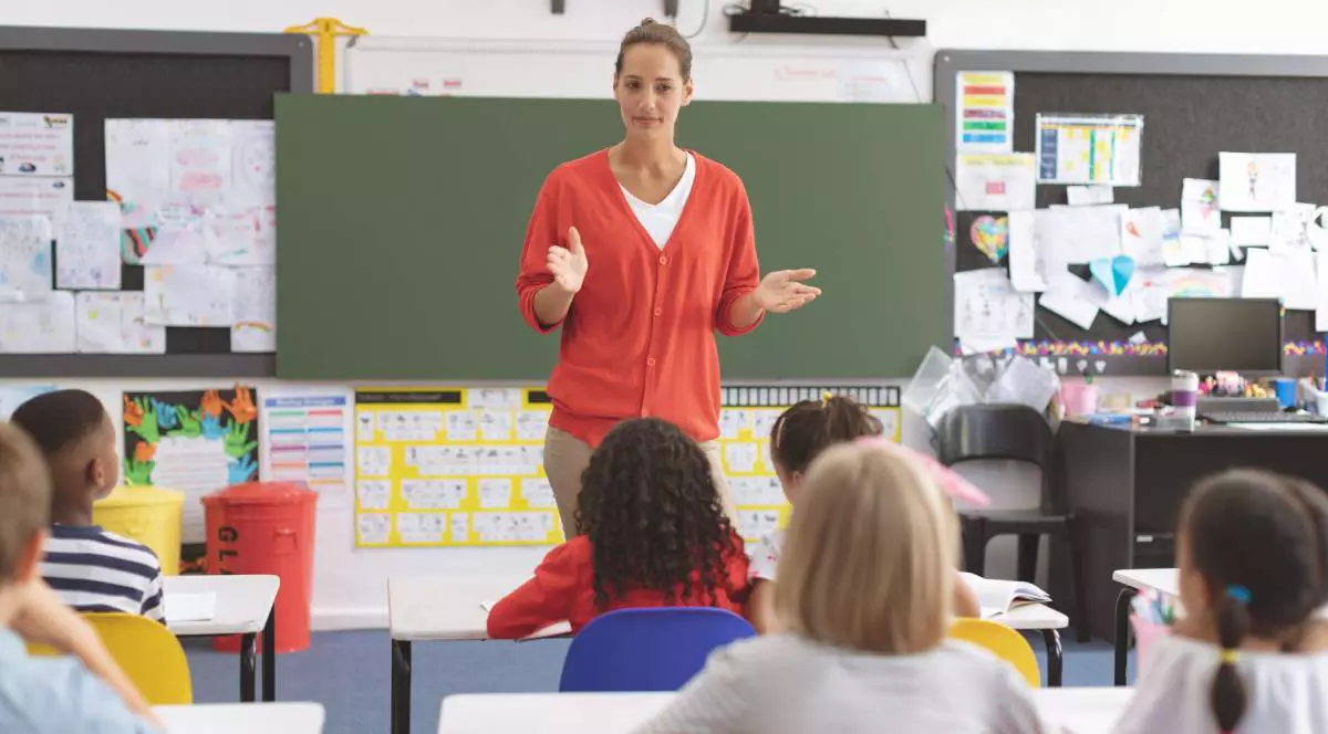 O profesoara in sala de clasa vorbește cu elevii din școala primara și le pregătește lecția, copiii sunt atenți la ceea ce le spune învățătoarea care stă în fața tablei