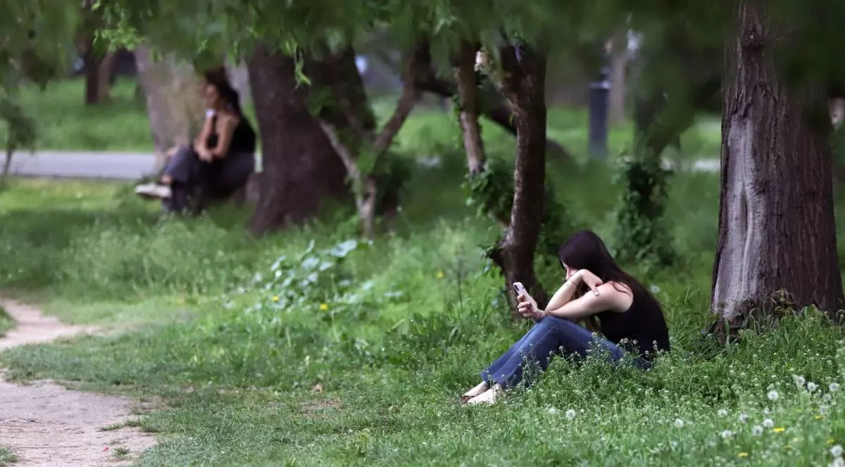 O fată cu maiou negru stă pe iarbă într-un parc și citește pe telefon, iar lângă ea se văd în spate doi tineri pe o bancă, București, parcul Cișmigiu, 25 aprilie 2025
