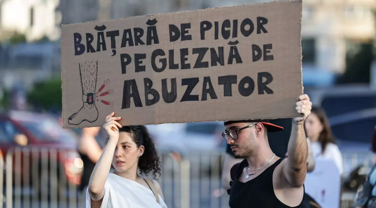 o femeie și un bărbat purtând un banner din carton maro pe care scrie „Brățară de picior pe gleznă de abuzator” în timpul protestului împotriva violenței de gen organizat de Centrul FILIA, 18 iunie 2025