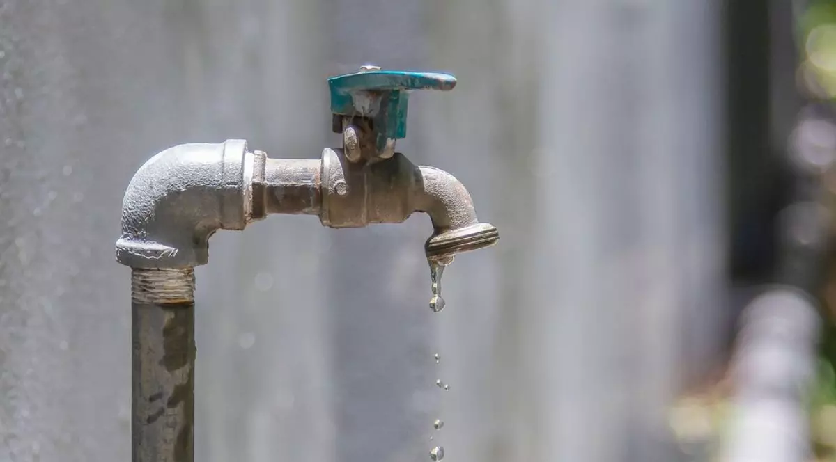 Robinet vechi din metal care picură apă în grădină, pe fundal un zid de ciment și natură