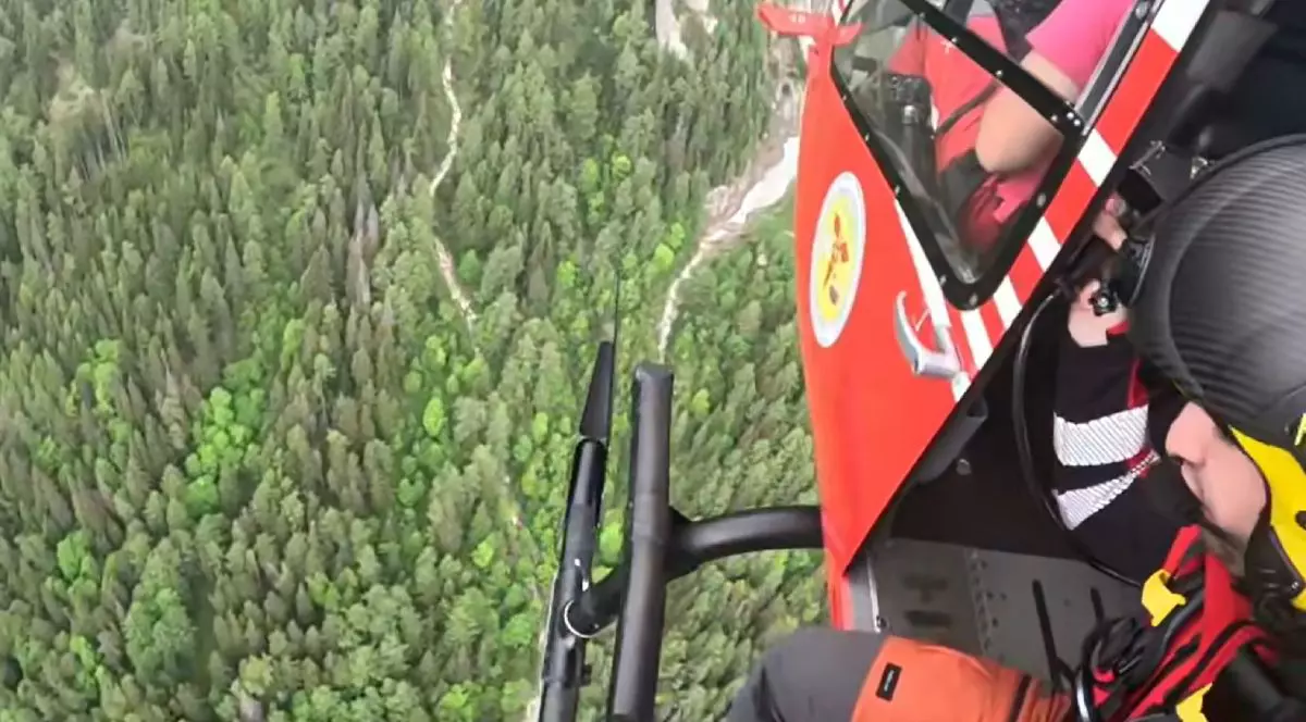 Imagini din elicopter cu salvamontist imbracat in rosu care se pregateste de coborare pe troliu pentru o operațiune de salvare deasupra unei paduri verzi