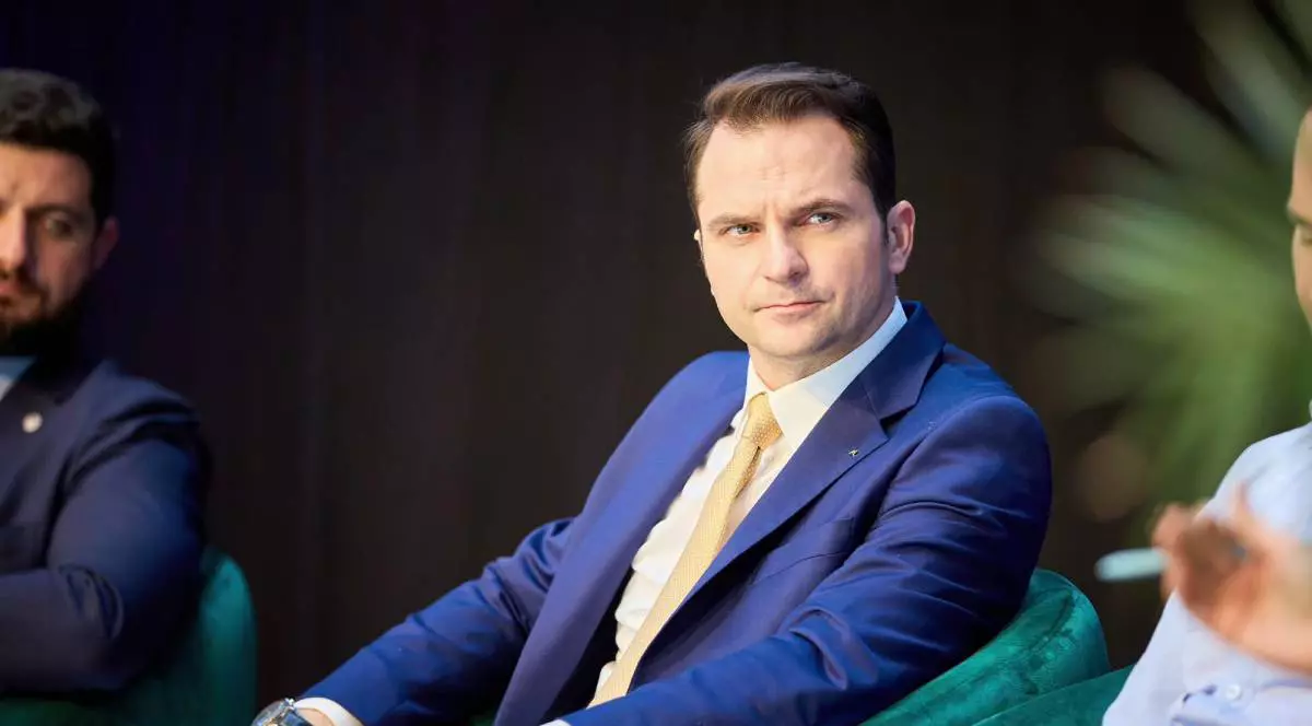 Ministrul Energiei, Sebastian Burduja, stând pe scaun în timpul unei conferințe, poartă un costum albastru și o cravată galbenă