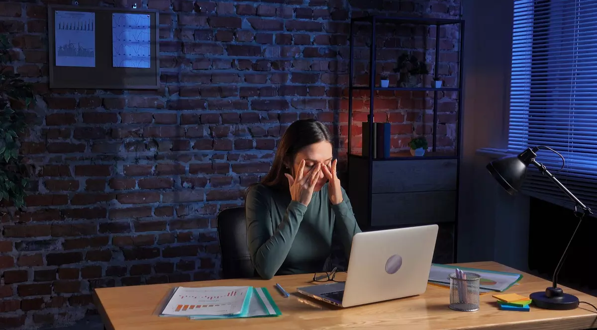 Femeie care lucreaza la laptop. Se afla la birou, in jur are rapoarte si pare ca o doare capul