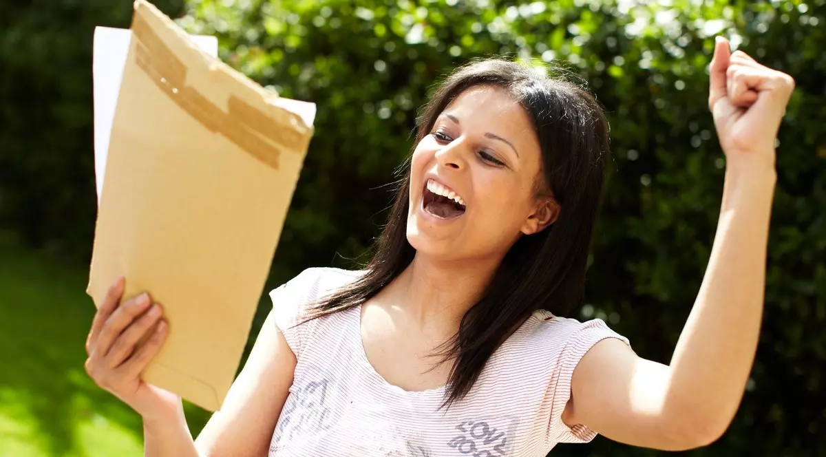 Poză cu o tânără care ține într-o mână un plic, iar cealaltă o are ridicată. Este surprinsă zâmbind