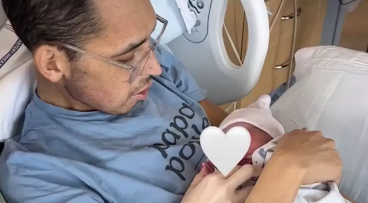 În imagine Tanner Martin, pe patul de spital, intr-un tricou gri petrol si cu ochelari, își tine în brate bebelusul nou născut, care are fata acoperita de o inimioara, 2024