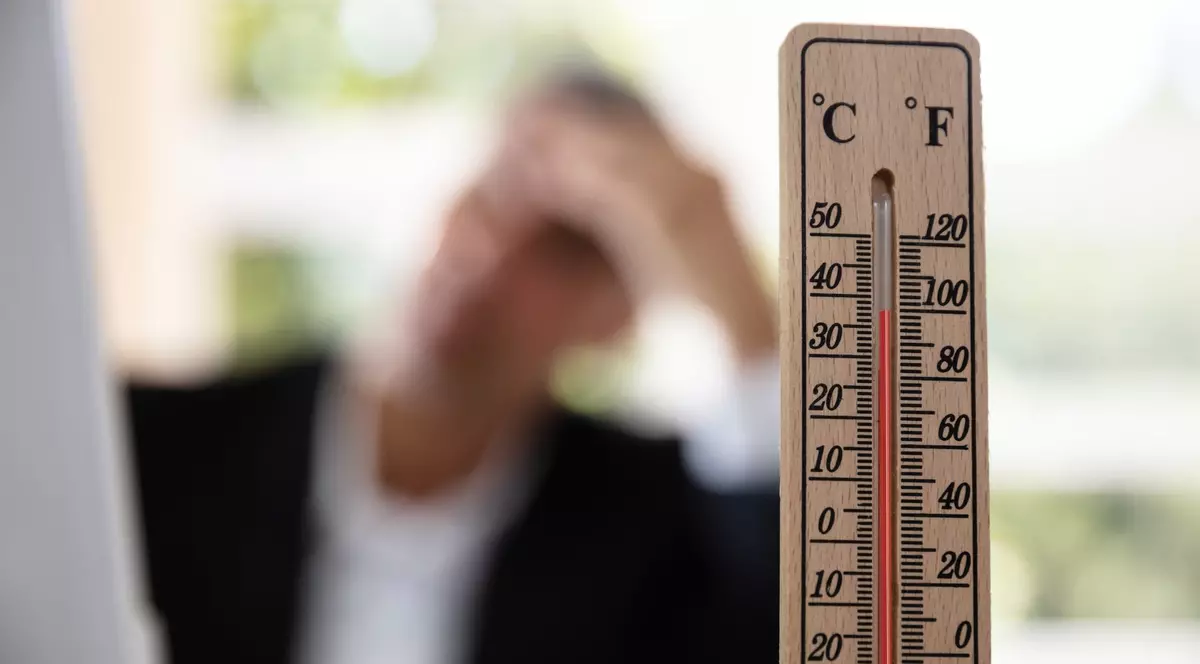 Imagine cu un termometru din lemn, iar în spate se află un bărbat la birou care are o mână pe frunte