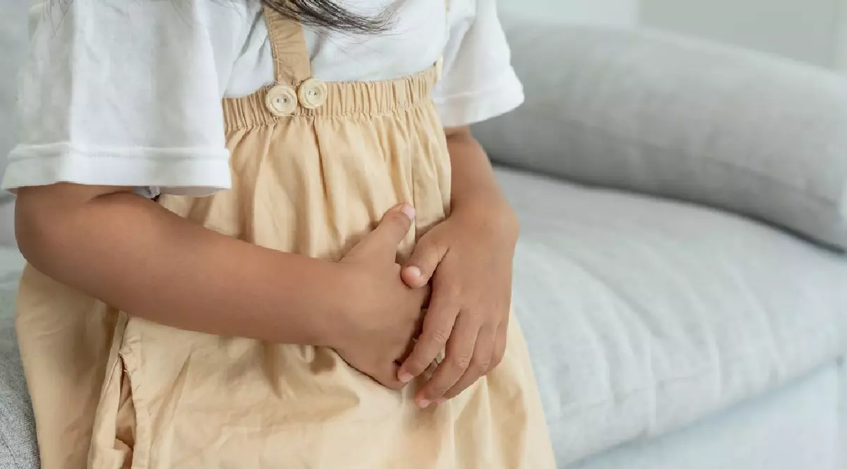 O fetiță, îmbrăcată în tricou alb și rochiță galbenă, stă pe o canapea gri și se ține cu mâinile de burtică