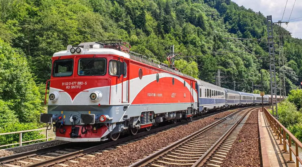 Un tren de călători cu locomotivă roșie merge pe calea ferată din România într-o zonă montană, este înconjurată linia de cale ferată de arbori cu frunze verzi.