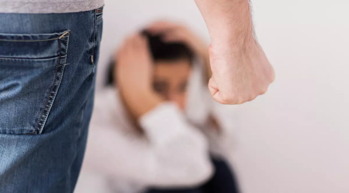 Imagine cu o femeie care stă pe podea și-și ține mâinile pe cap, iar în stânga se află un bărbat cu pumnul încleștat