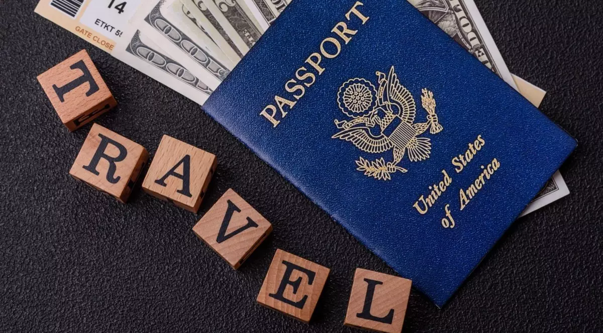 pașaport american cu coperta albastră în care se află mai multe bancnote de 100 de dolari, așezat pe o masă neagră, alături de șase cuburi de lemn pe care sunt inscripționate literele T R A V E L