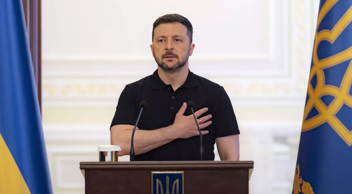 Voldimir Zelenski, îmbrăcat într-un tricou polo cu mânecă scurtă, cu mâna stângă la piept, stând la un pupitru de lemn închis la culoare pe care sunt amplasate două microfoane
