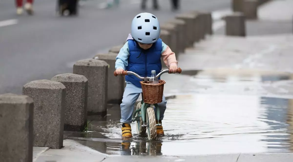 Un băiețel cu cască bleu, îmbrăcat cu mânecă lungă și vestă, este pe bicicletă și trece printr-o baltă de pe trotuar. Are adidași galbeni, iar în coșul de paie pus în fața bicicletei este o sticlă cu apă