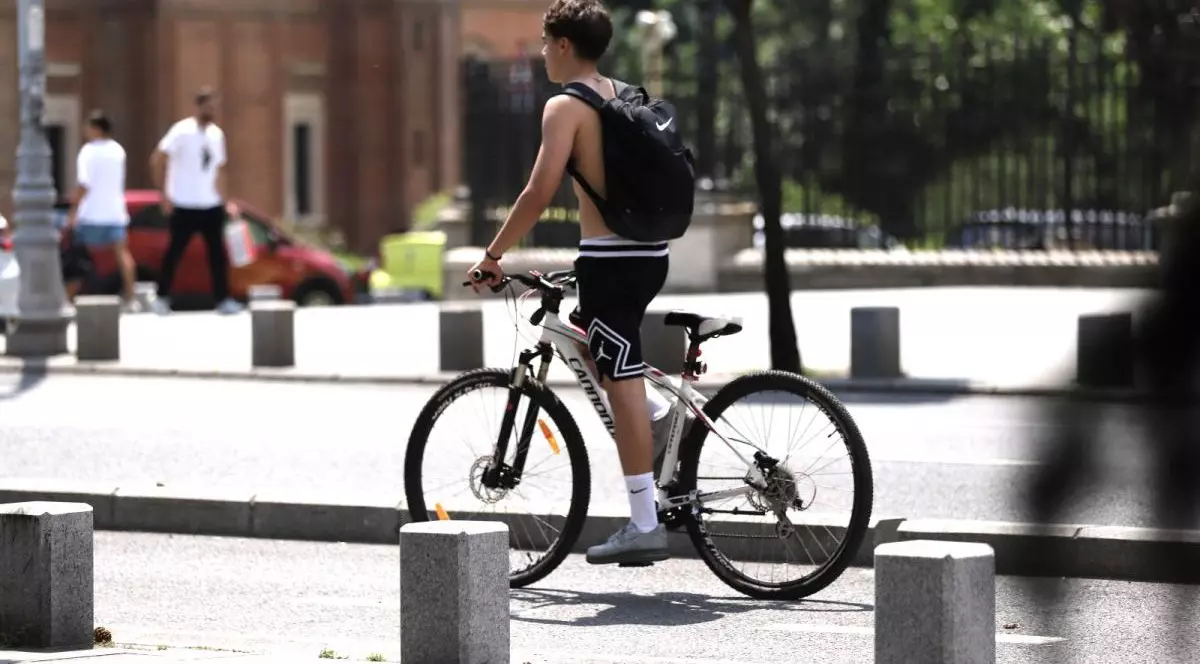 Un băiat fără tricou și cu un ghiozdan negru în spate merge pe bicicletă în picioare, într-o zi caldă de vară. În stânga se văd doi oameni în tricouri albe, iar în stânga o zonă verde dintr-un parc
