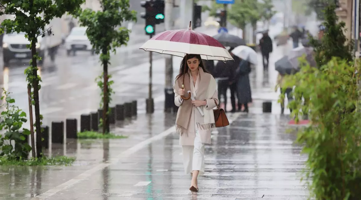O tânără îmbrăcată în culori deschise și pantofi cu toc, ținând o umbrelă deschisă în mână, merge pe stradă într-o zi cu ploaie, iar în spate se vede un semafor la culoarea verde