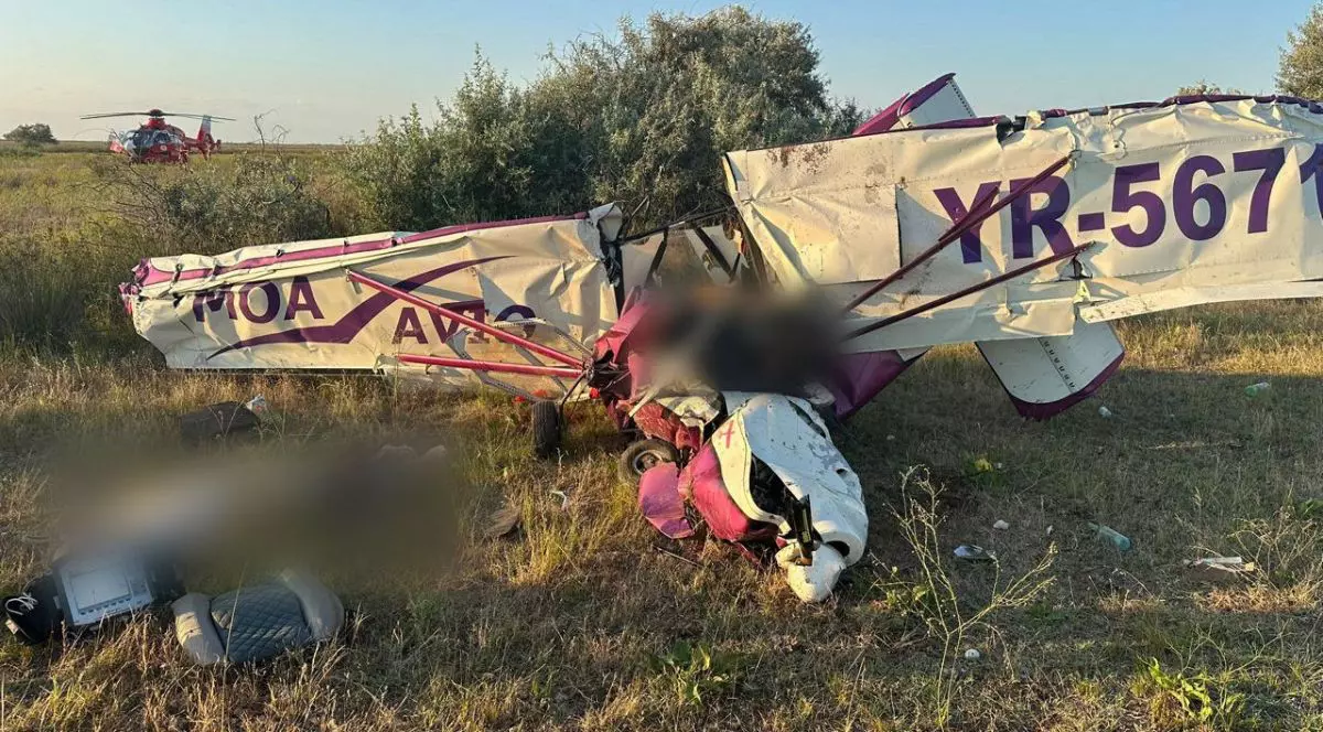 un avion alb de mici dimensiuni, cu detalii roz, prăbușit la pământ, cu aripile deteriorate. lânga epava avionului se pot observa scaunele avionului, rupte și aruncate la o distanța de un metru