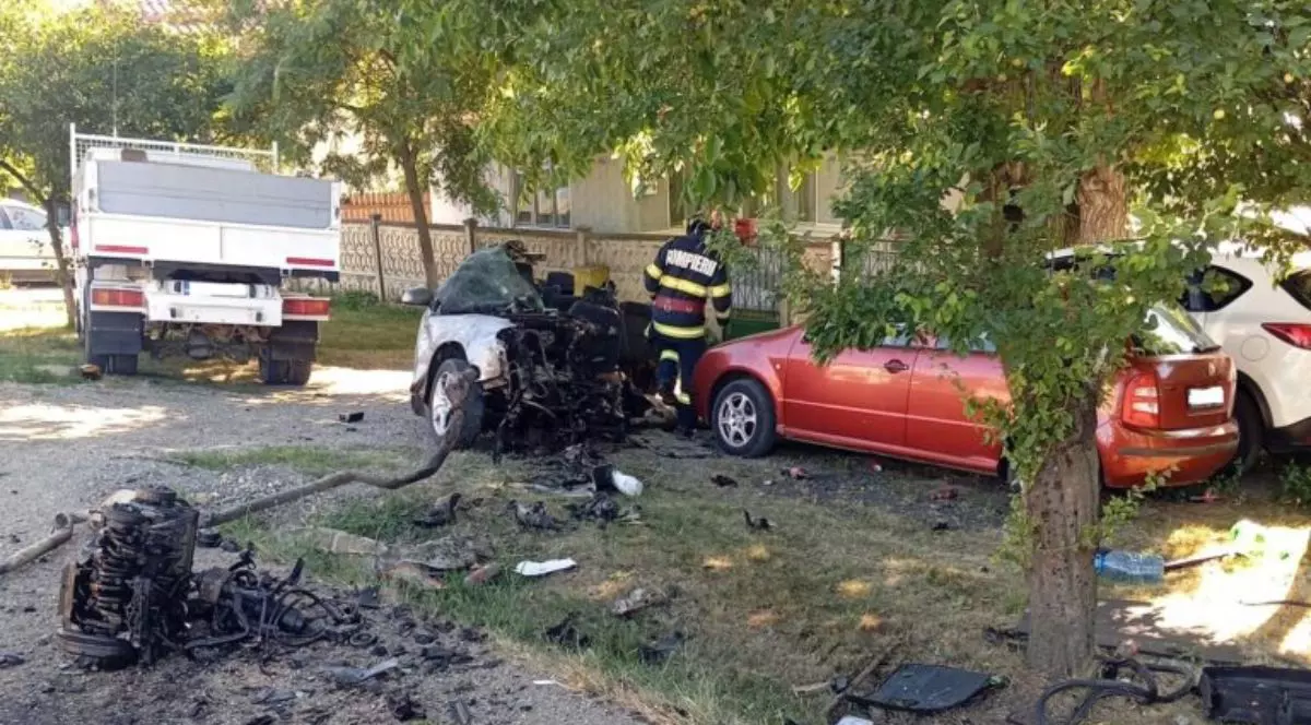 un autoturism alb, complet distrus în partea din față, cu motorul și alte componente aruncate la câțiva metri distanță de caroserie. Lângă autoturism se află un pompier îmbrăcat în uniformă roșie și pe marginea drumului, sunt parcate alte două mașini, una roșie și una albă, care par să fi fost avariate ușor.