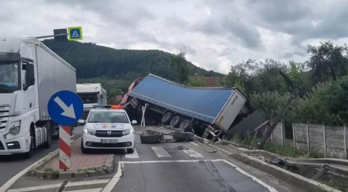 În prim-plan, în mijlocul drumului, se află o mașină de poliție cu lumini roșii aprinse. În spatele ei, un camion de mari dimensiuni cu remorcă albastră este răsturnat pe marginea drumului, avariat, cu partea din spate căzută într-o zonă joasă lângă un gard de beton. Pe carosabil sunt vizibile roți desprinse și resturi împrăștiate. În partea stângă a imaginii, alte camioane sunt oprite
