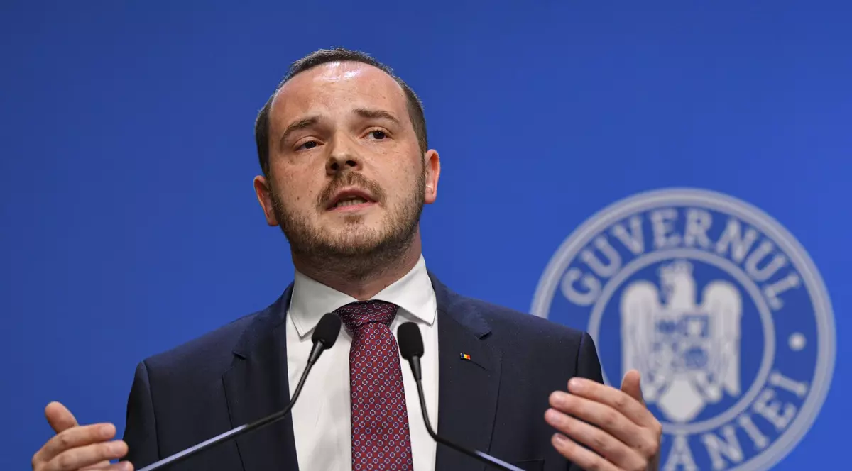 Alexandru Rogobete surprins în timpul unei conferințe de presă la Guvern vorbind și având mâinile ridicate. Poartă un sacou negru, o cămașă albă și o cravată grena
