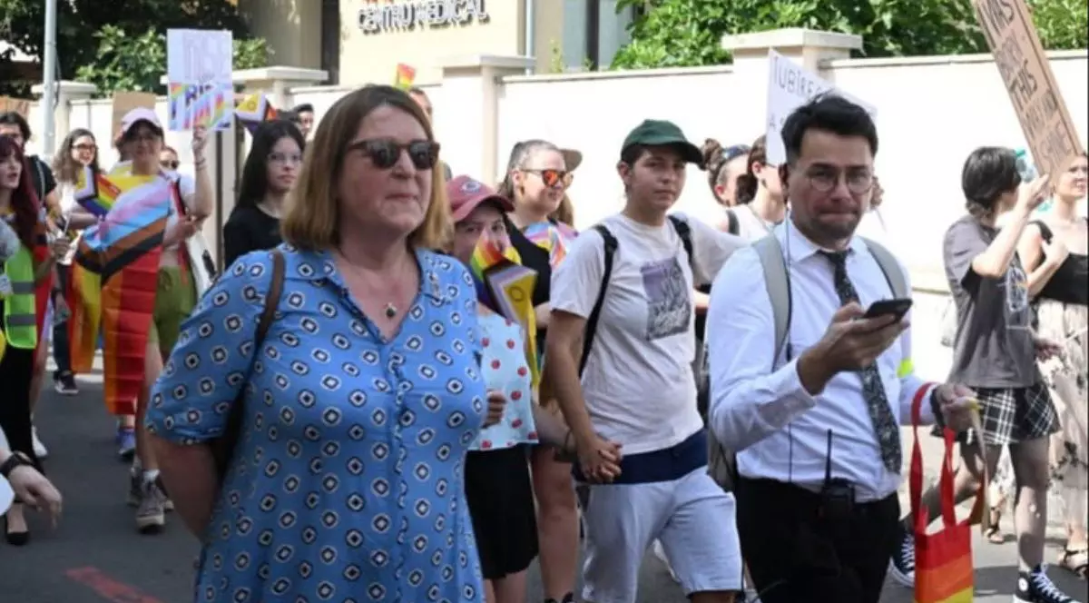 În imagine ambasadoarea Olandei, Willemijn van Haaften, într-o richie bleu, cu ochelari de soare si mainile la spate, participă la Oradea Pride, alături de activistul Victor Ciobotaru, in comasă albă si pantalon negru, in jurul lor mai mult persoane care participa la parada, unele tin pancarde. 26 iulie 2025, Oradea