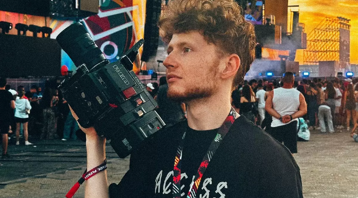 un băiat la un concert pe plajă cu o camera pe umeri și mulți oameni in spate