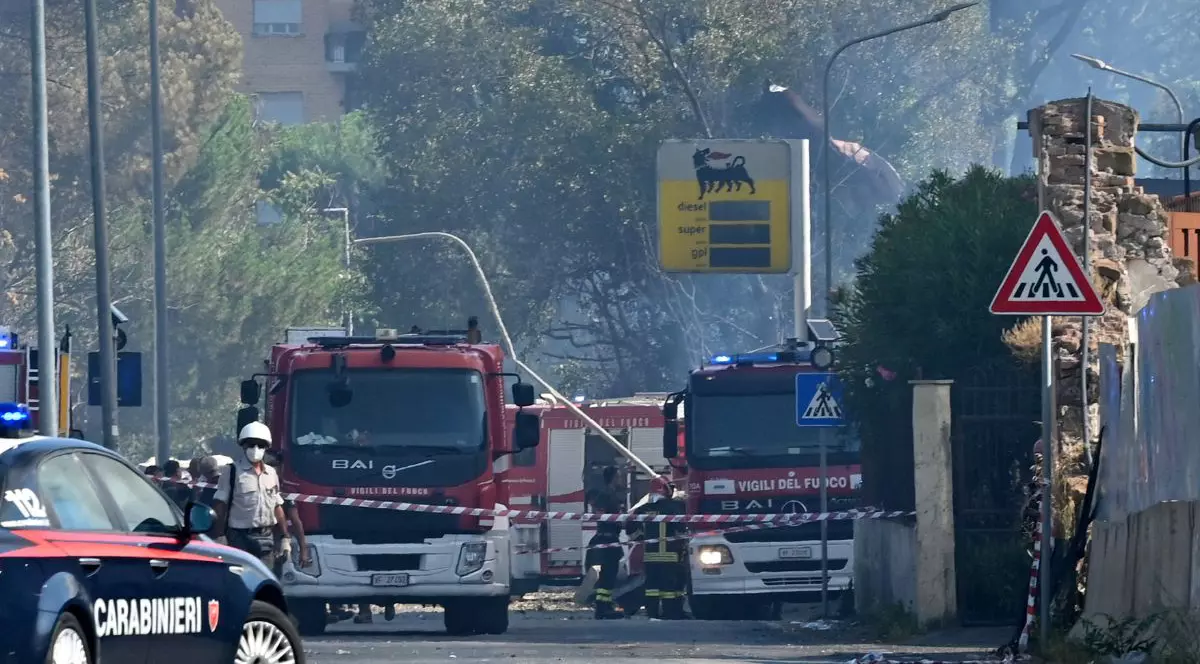 Două autospeciale de stingere a focului sunt staționate în spatele unei benzi colorate în dungi roșii și albe care delimitează zona de pericol. în partea dreaptă a fotografiei se poate observa semnul luminos al unei benzinării ENI, care înfățișează un câine negru pe un fundal galben. în dreapta imaginii se află o mașină neagră de poliție, cu textul „CARABINIERI” în font gros și alb, pe portierele mașinii