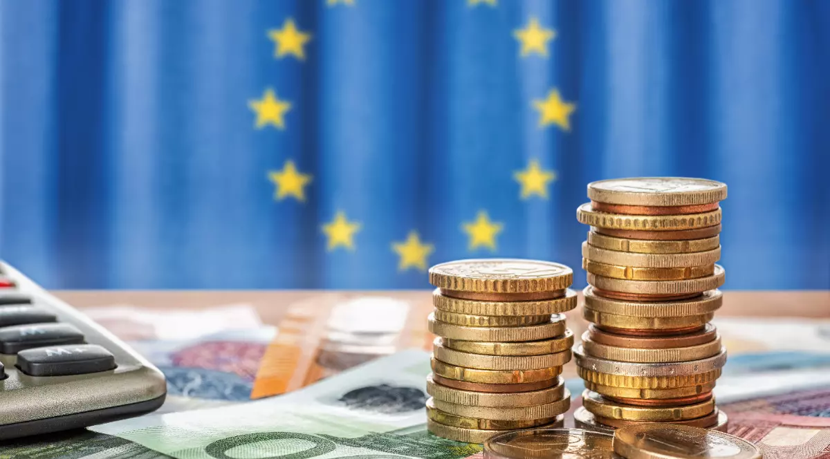Monede așezate unele peste altele, bancnote, un calculator, iar în fundal este steagul Uniunii Europene