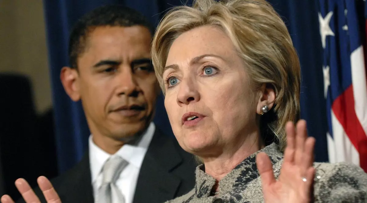 Barack Obama, in plan indepartat, intr-un costum negru, camasa alba si cravata, asculta discursul lui Hillary Clinton aflata in prim-plan. In spate se observa drapelul SUA