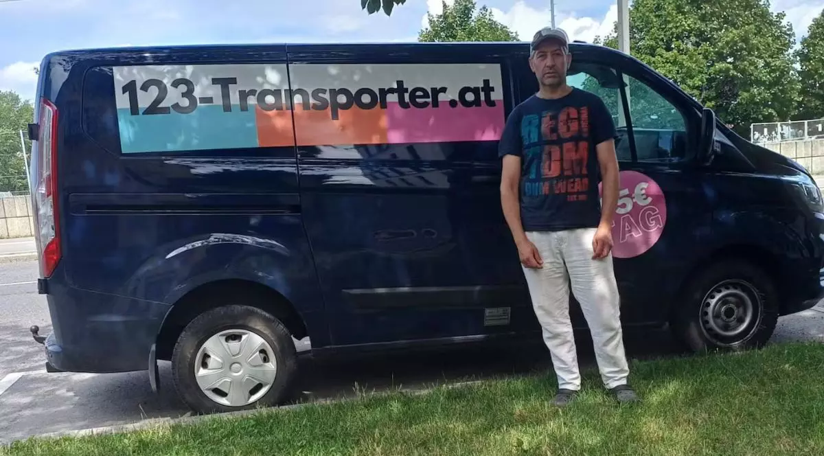 un bărbat îmbrăcat într-un tricou negru cu text în degrade albastru și portocaliu, pantaloni albi și cu o șapcă neagră pe cap, stând lângă o dubă Ford Custom bleumarin cu un colant alb, cu dungi albastre, portocalii și roz, pe care este imprimat textul 123-Transporter.at