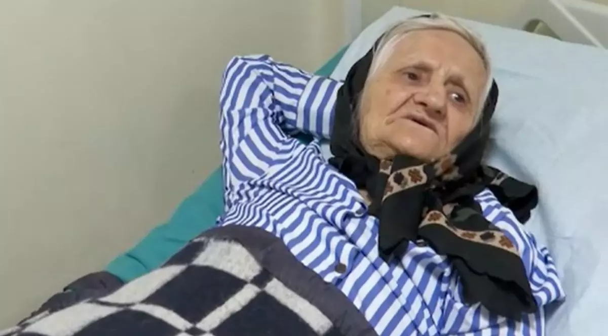 Bătrâna din Iași, pe patul de spital, cu un batic negru pe cap, îmbrăcată cu o bluză de pijama în dungi alb-albastre