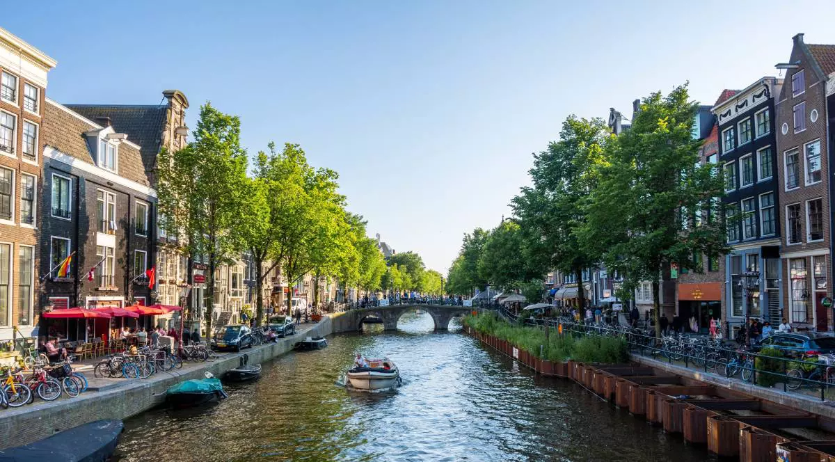 Unul dintre canalele din Amsterdam copacii din jur au frunzele verzi, bicicletele sunt parcate pe marginea apei