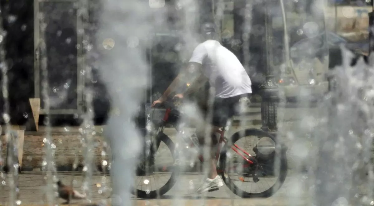 Un tânăr pe bicicletă, fotografiat cu spatele, în tricou alb și cu cască pe cap, trece prin dreptul unor fântâni din București, într-o zi caniculară de vară, București, 17 iulie 2025.
