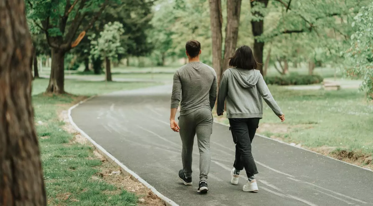 O fata si un baiat merg pe jos pe o alee din parc, in jurul lor sunt multi copaci verzi