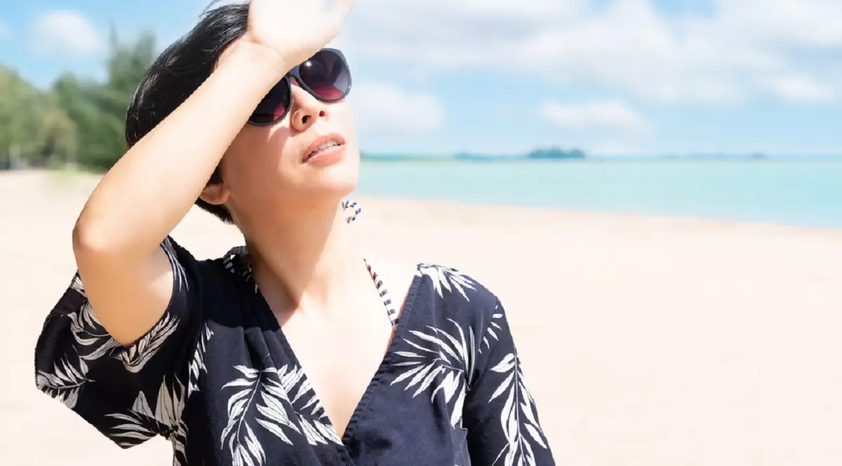 Ce este radiaţia ultravioletă şi cum interpretăm indicele UV - O femeie brunetă se află pe plajă, la malul mării, într-o ținută ușoară de plajă și cu ochelari, se uită spre soare, ținându-și mână în dreptul ochiilor