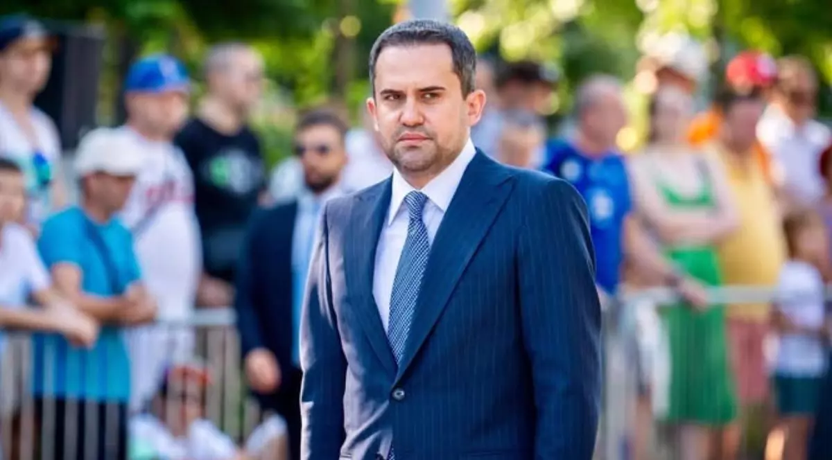 Ministrul Ciprian Șerban, în costum albastru si cu cravata, cu cămasă albă, surprins in aer liber, vara, in spatele lui se vede o multime de oameni
