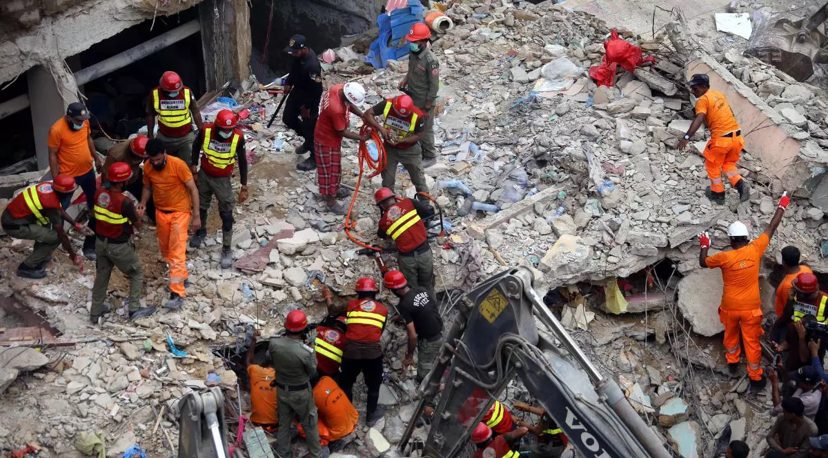 dărâmături – bucăți mari de beton, fier beton, praf și resturi de mobilier. printre ruine se află peste 15 persoane, majoritatea purtând căști de protecție roșii sau albe și veste reflectorizante. În partea de jos a imaginii, se vede brațul unui excavator.