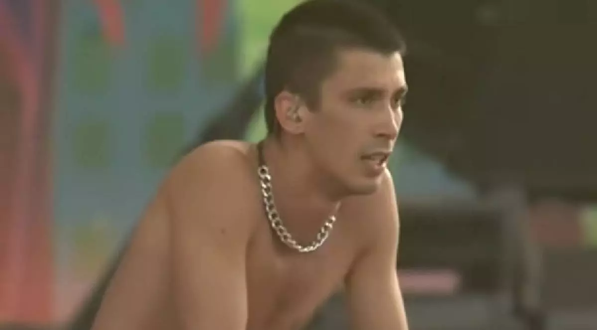 În imagine, artistul Cojo stă pe scenă privindu-și fanii. Este la bustul gol și în mână ține un microfon