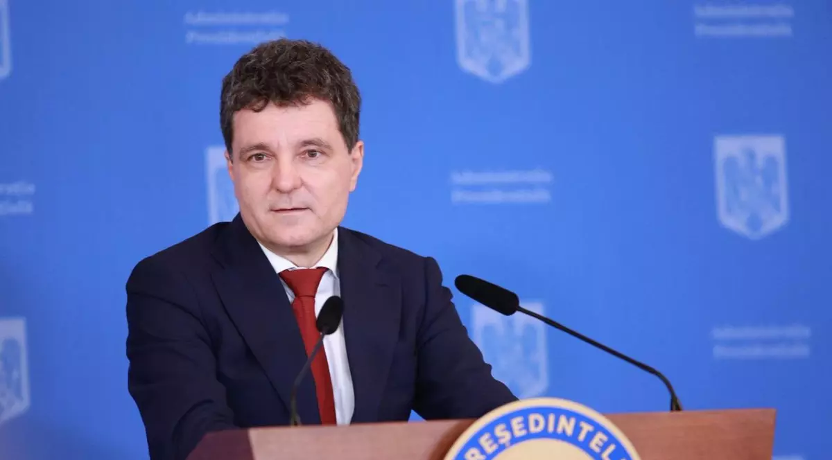 Nicușor Dan, îmbrăcat într-un sacou negru, o cămașă albă și o cravată roșie, în timpul unei conferințe de presă, vorbind la microfon în fața unui pupitru cu stema administrației prezidențiale