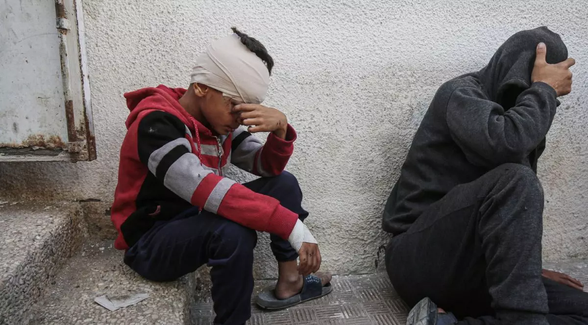 Un copil palestinian lovit si bandajat la cap, cu mana stanga la ochi, sta asezat pe niste trepte din betan, langa el un barbat in haine ponosite, gri, cu mana dreapta la cap si fata acoperita cu glugă. 26 mai 2925