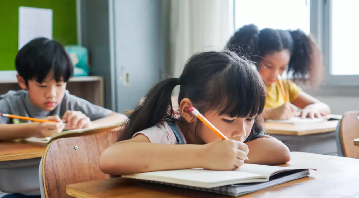Elevă stând în bancă și scriind în caiet cu creionul, studiind, educație, învățare. Copii asiatici în clasă. Diversitate în rândul elevilor.