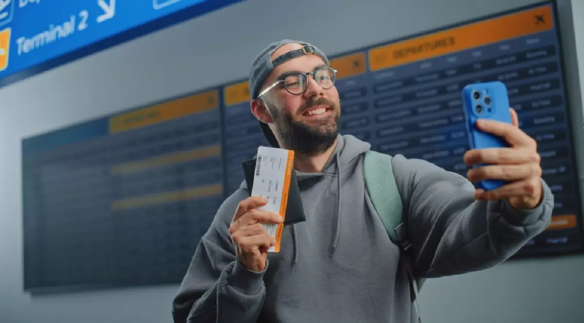 Cum să găsești bilete de avion ieftine - Un bărbat, care poartă o șapcă pe cap și ochelari de vedere, își face un selfie, într-un aeroport, ținând bucuros un bilet în mâna dreapta