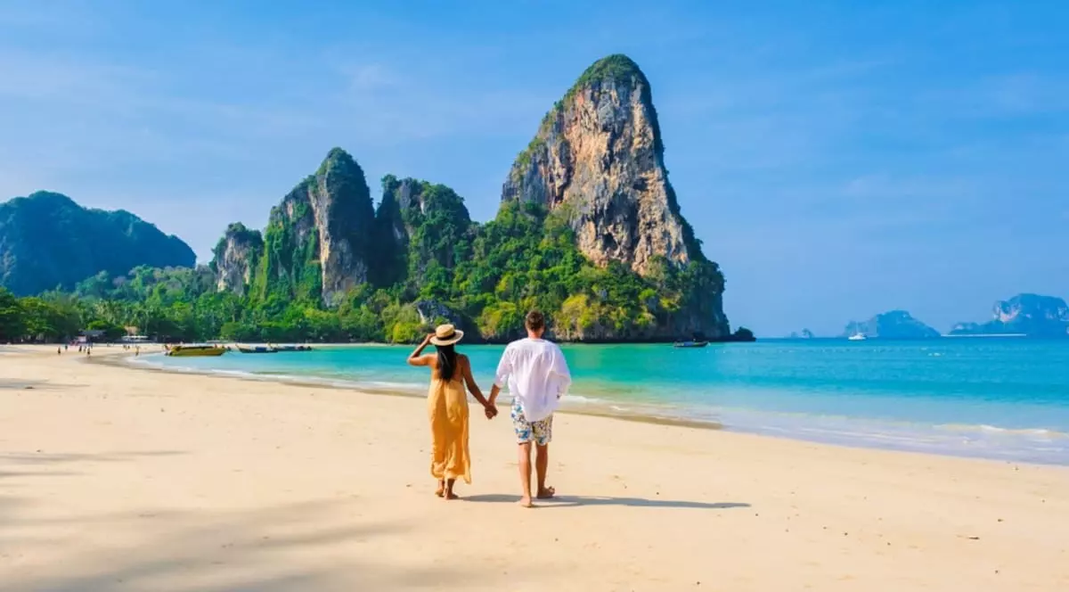 Doi tineri merg pe o plajă spre o stâncă împădurită care intră în mare