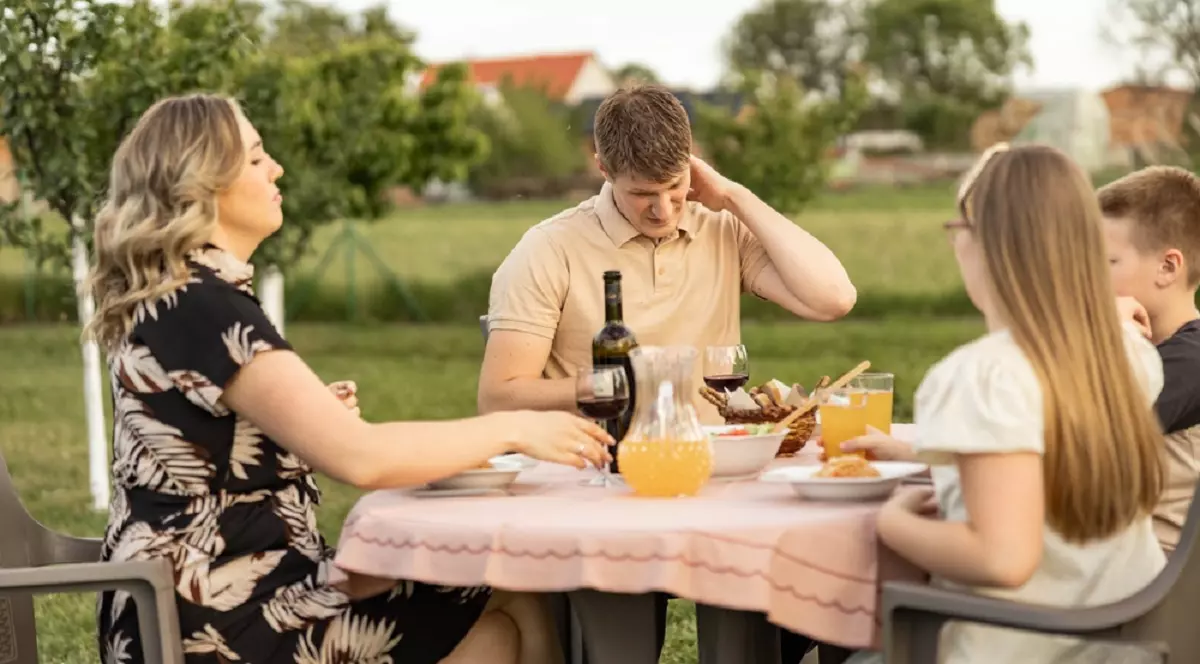 Cum scapi de ţânţari fără a folosi insecticid - O familie cu doi copii se bucură de o masă în grădină, însă tatăl este deranjat de mușcăturile de țânțari și este surprins în timp ce se scarpină pe ceafă