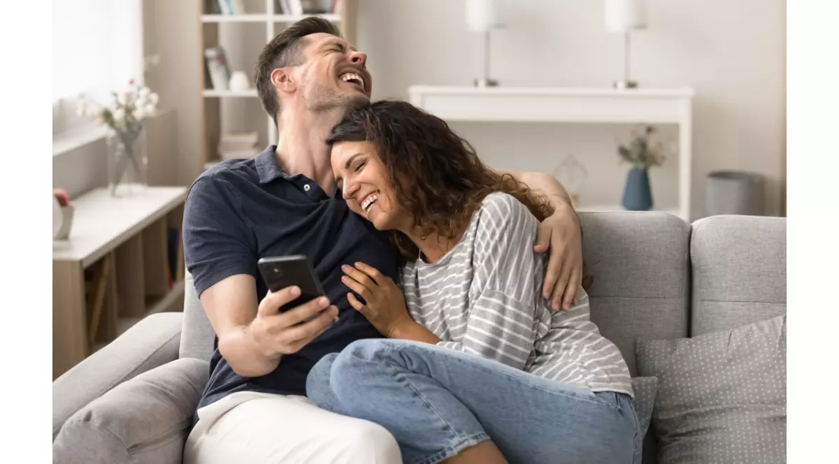 Cuplu care stau imbratisati pe canapea in sufragerie. Rad la ceva de pe telefon
