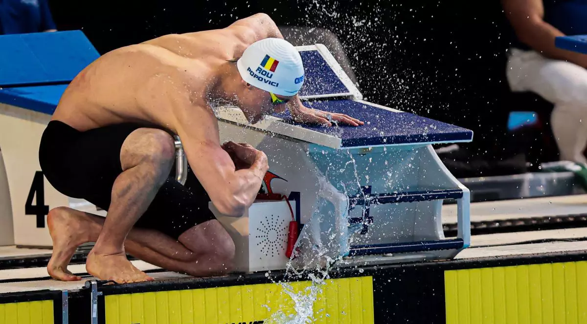 Înotătorul român David Popovici, cu casca alba si sort, la sfartul unuei competitii, la marginea bazinului de înot.